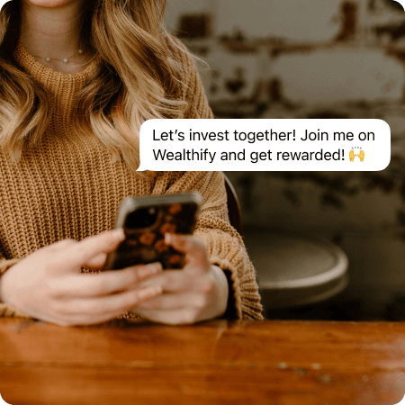 A woman sat at a table looking at her phone. Superimposed on the image shows a notification that reads: 'Let's invest together! Join me on Wealthify and get rewarded!'